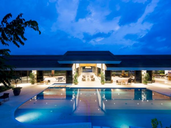 A modern building at dusk with a large, illuminated swimming pool in the foreground, surrounded by seating.