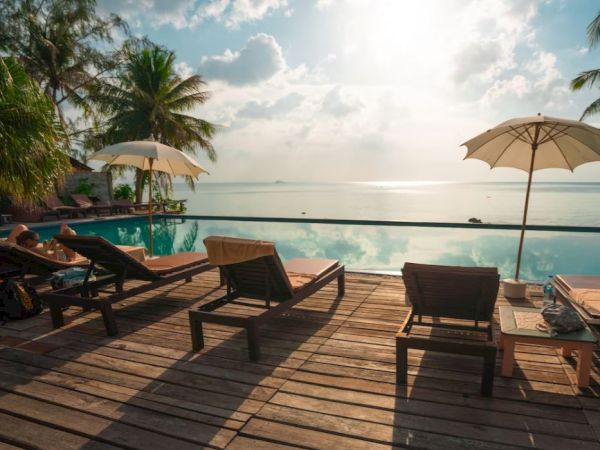 A serene poolside scene with lounge chairs, umbrellas, palm trees, and an ocean view under a partly cloudy sky.