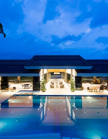 A modern building with large open spaces is seen at night, featuring a brightly lit swimming pool and seating area under a cloudy blue sky.