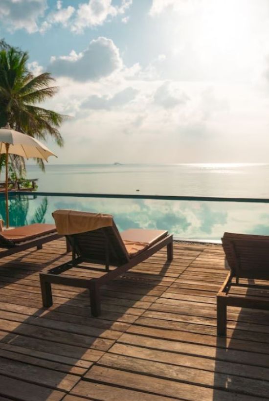 The image shows a serene poolside setting with wooden lounge chairs, umbrellas, palm trees, and a view of the ocean and sky.