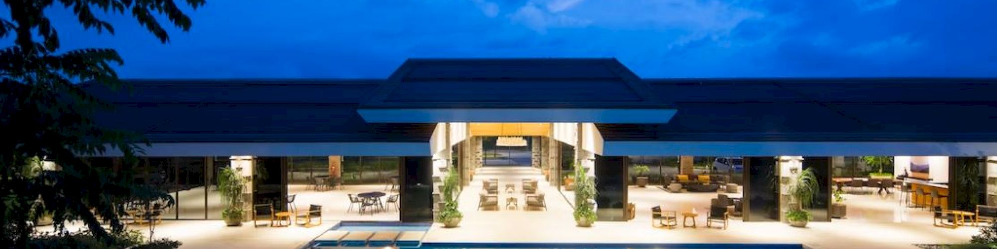 A luxurious poolside with a modern building illuminated at night, featuring lounge chairs and a serene ambiance, under a vivid blue sky.