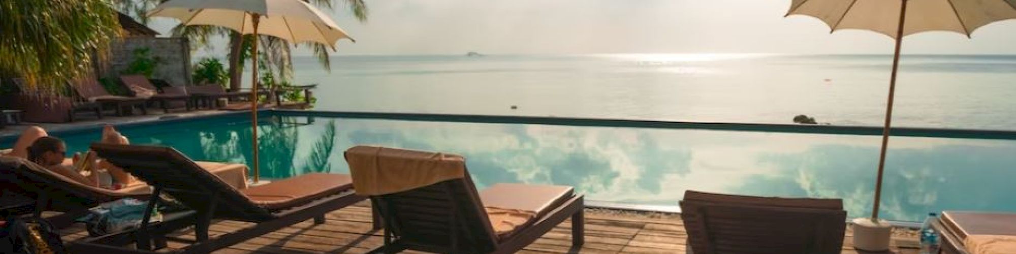 A serene poolside setting with loungers, umbrellas, and palm trees overlooks a calm ocean under a partly cloudy sky.