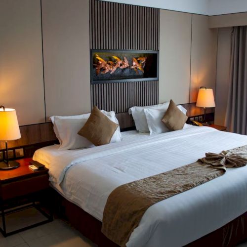 The image shows a hotel room with a large bed, decorative pillows, lamps, and a small wall fireplace, creating a cozy atmosphere.