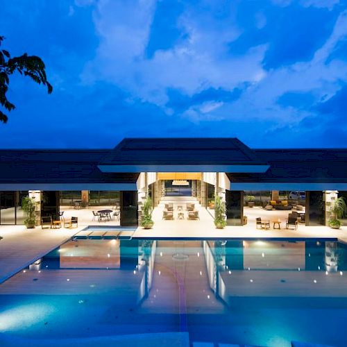An illuminated modern building with a large pool in front, surrounded by plants, set against a blue evening sky.