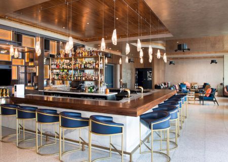 A modern bar with a central counter, stylish lighting, blue stools, and shelves stocked with various bottles, creating a cozy atmosphere.