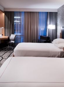 The image shows a modern hotel room with two beds, a wall-mounted TV, a work desk, and ambient lighting.