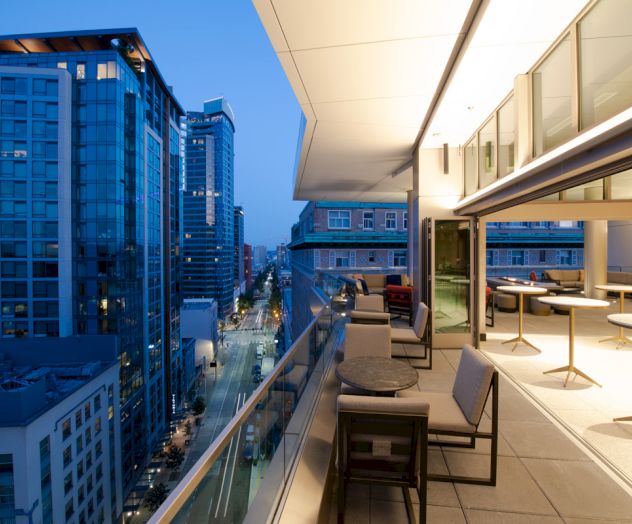 A modern rooftop patio with tables and chairs overlooks a city street with tall buildings under a twilight sky.