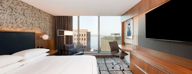 A modern hotel room features a large bed, wall-mounted TV, desk, and window with a city view, creating a cozy, elegant atmosphere.
