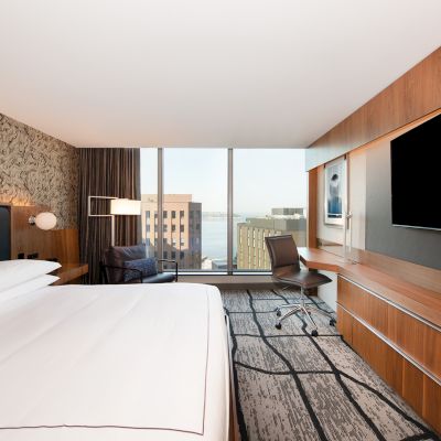 A modern hotel room with a large bed, desk, chair, TV, and window offering a city view.