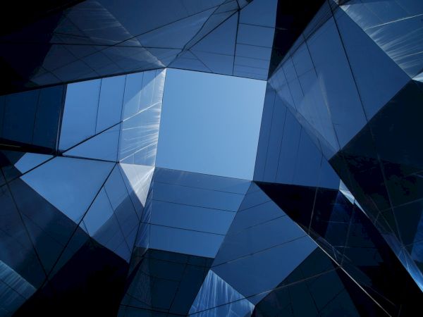 Geometric architectural structures reflect the sky, creating intricate patterns of glass and metal with a blue background.