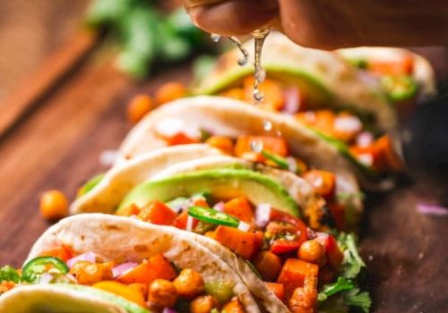 A hand squeezes lime over a row of tacos filled with chickpeas, vegetables, and garnished with cilantro on a wooden board.