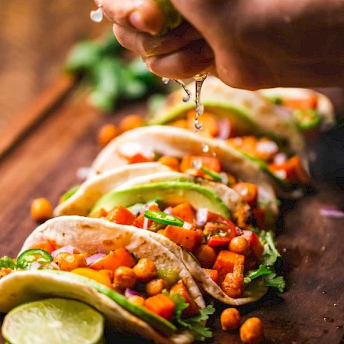 A hand squeezes lime over a row of tacos filled with chickpeas, vegetables, and garnished with cilantro on a wooden board.