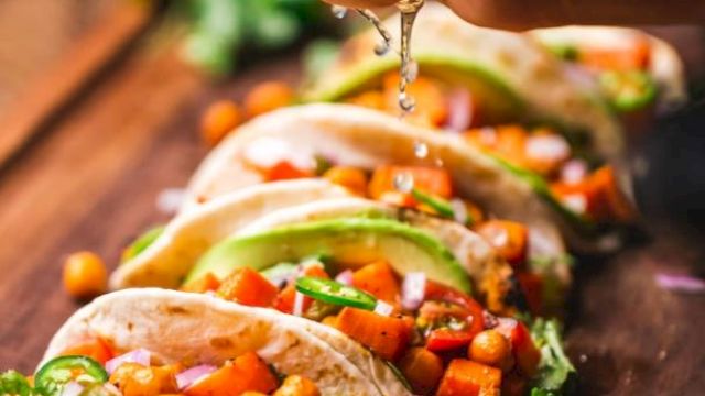 A hand squeezes lime over a row of tacos filled with chickpeas, vegetables, and garnished with cilantro on a wooden board.