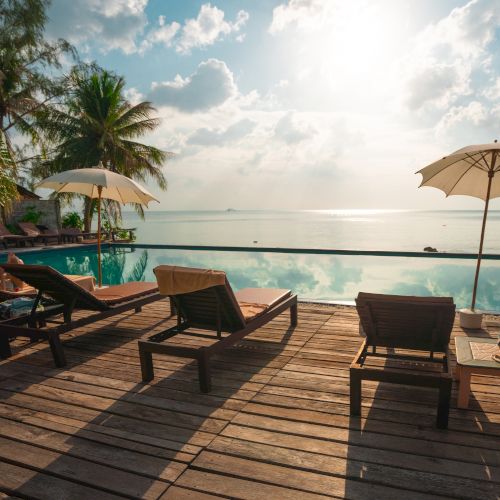 A tranquil tropical scene with sun loungers by a pool, under umbrellas, facing the ocean with palm trees and a sunlit sky.