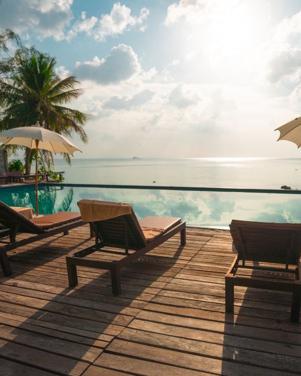 A tranquil tropical scene with sun loungers by a pool, under umbrellas, facing the ocean with palm trees and a sunlit sky.
