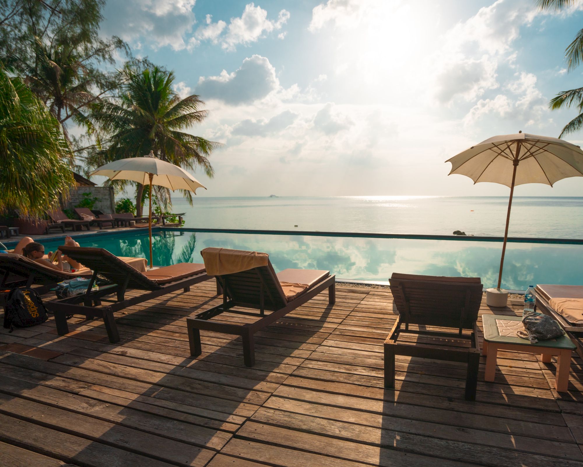 Lounge chairs and umbrellas by a pool overlook a beach scene, with palm trees and a clear sky in the background.