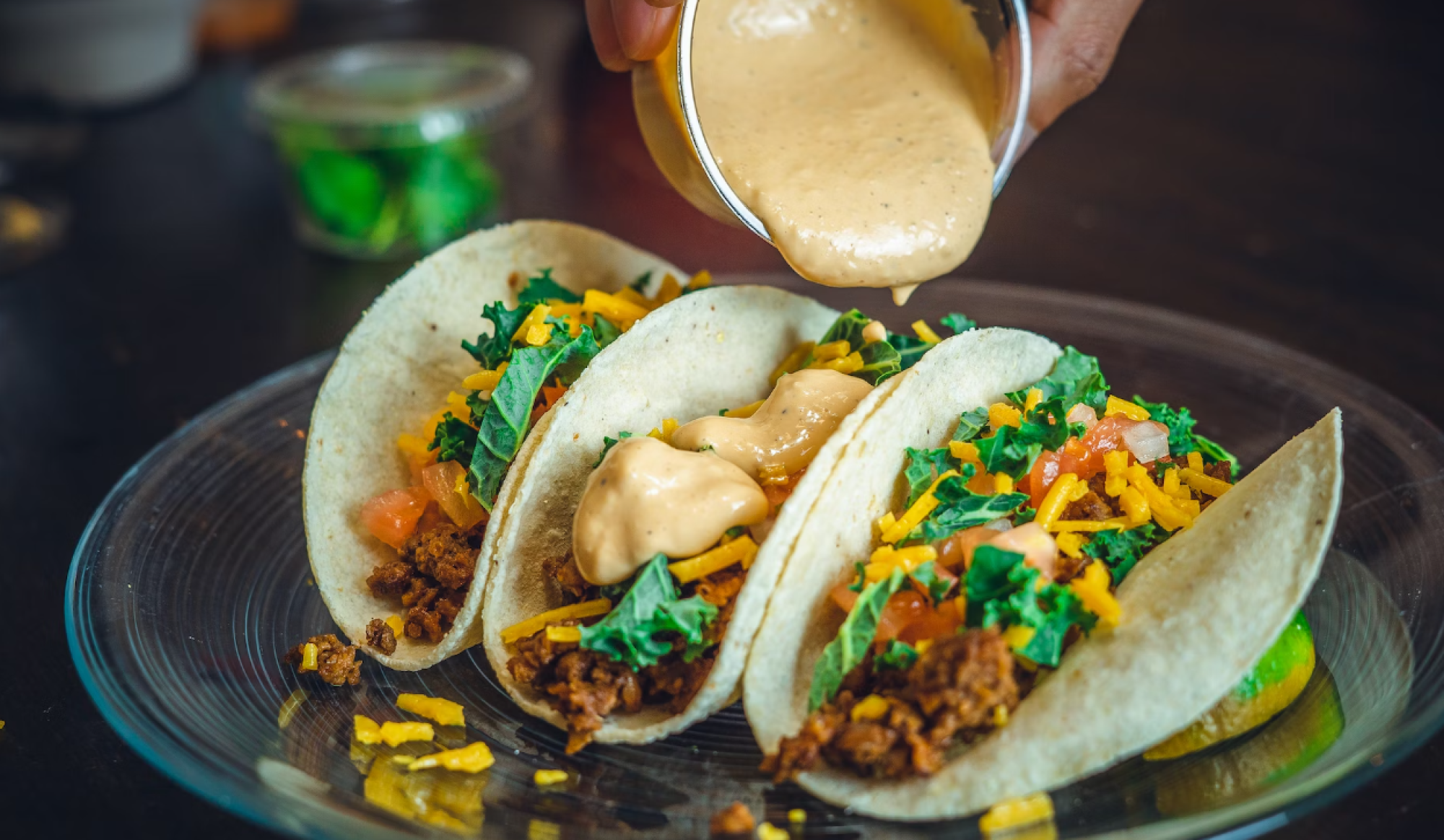 Three tacos filled with seasoned meat, lettuce, tomatoes, cheese, and sauce being poured over them on a plate.