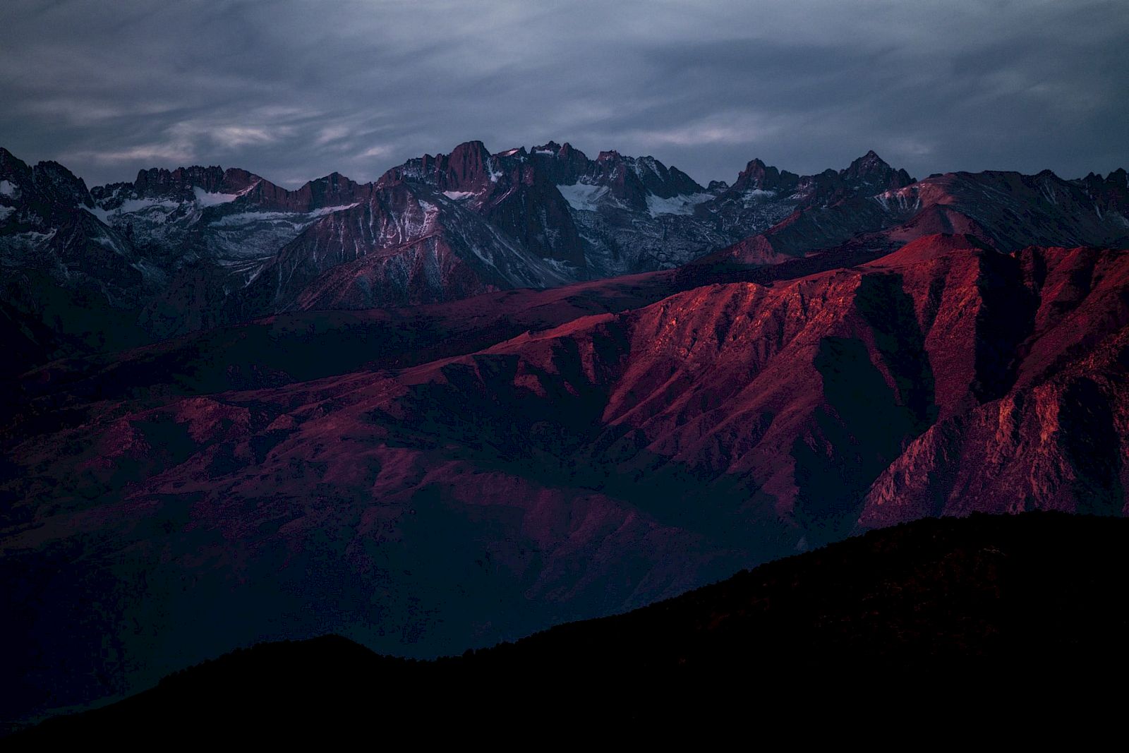 A mountain range at dusk with reddish hues on the peaks under a dramatic, cloudy sky.