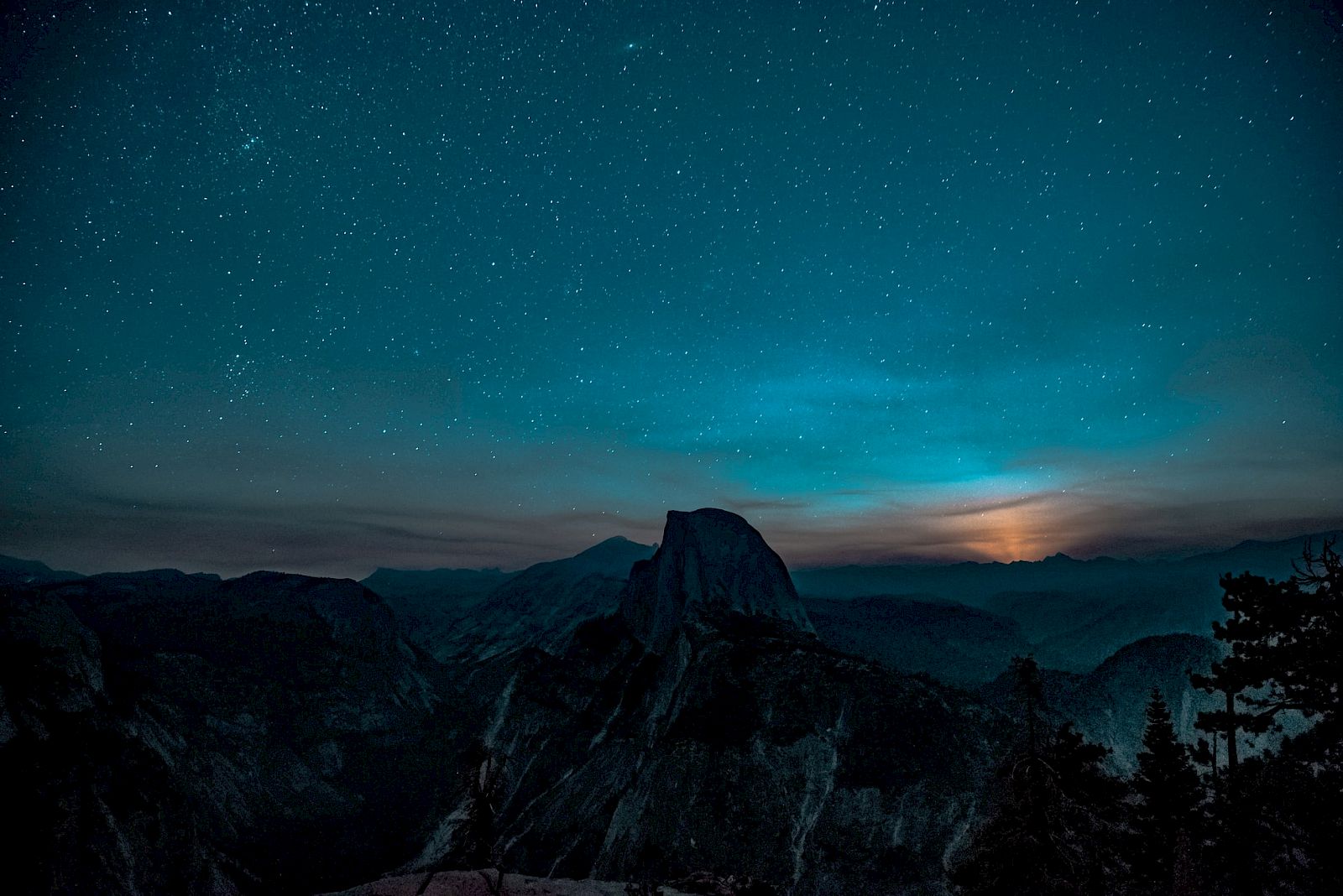A night sky filled with stars over a mountain landscape, with faint light illuminating the horizon behind the peaks.