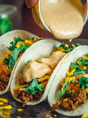 Three tacos filled with meat, cheese, tomatoes, lettuce, and sauce being poured on top; a small container with green contents in the background.