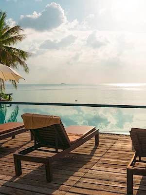 The image shows a serene beachside pool with loungers, umbrellas, and palm trees, offering a stunning view of the calm ocean and sky.
