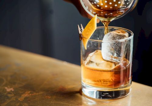 A cocktail being poured into a glass with ice and a twist of citrus peel on a bar counter.