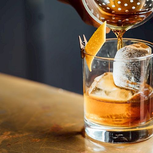 A cocktail being poured into a glass with ice and a twist of citrus peel on a bar counter.