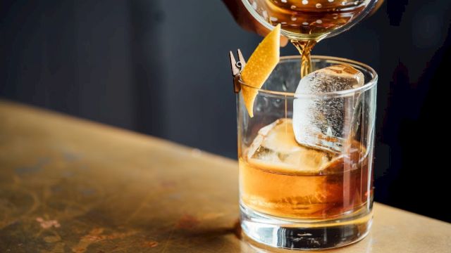 A cocktail being poured into a glass with ice and a twist of citrus peel on a bar counter.