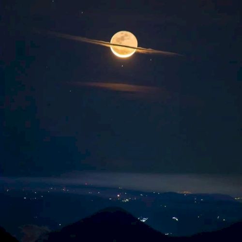 A bright full moon shines in the night sky, partially obscured by clouds, resembling Saturn with its rings, over a landscape with lights.