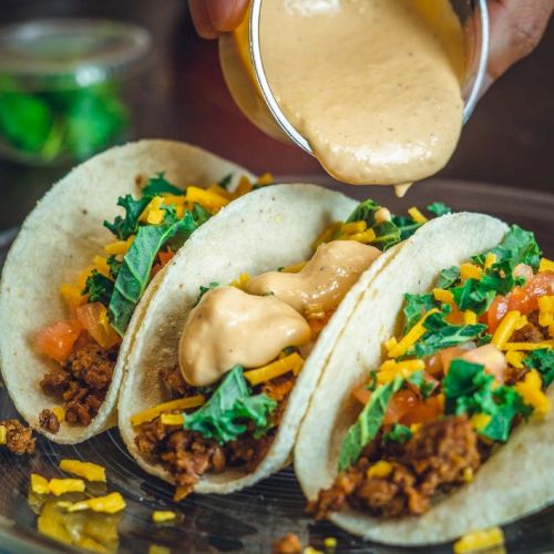 Three tacos filled with ground meat, cheese, lettuce, and tomatoes, topped with a creamy sauce being poured on them.