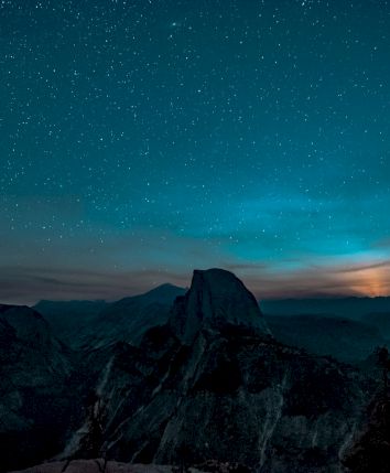 A starry night sky over a mountainous landscape with a prominent peak silhouetted, under a glowing horizon.