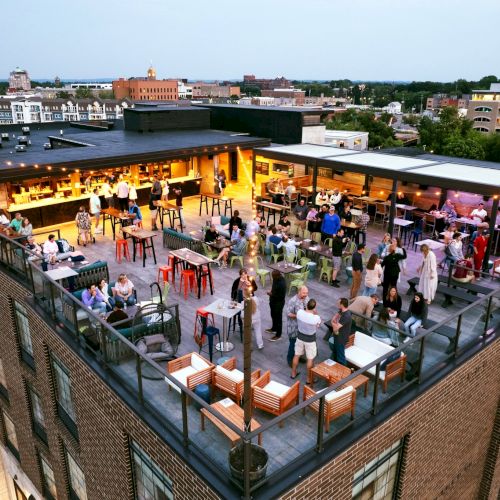 Rooftop bar scene with people socializing, seated at tables with drinks, while enjoying an elevated city view at dusk.