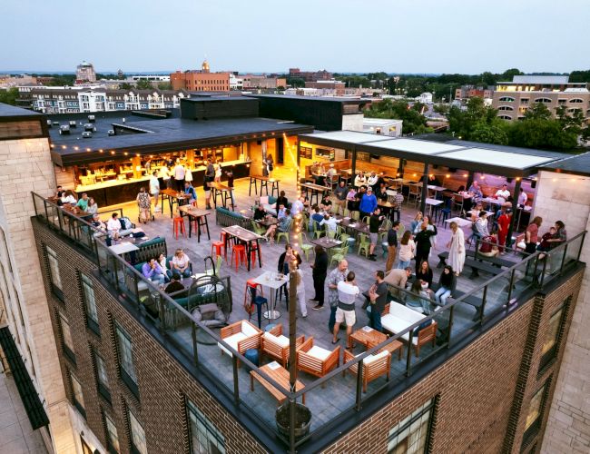 Rooftop bar scene with people socializing, seated at tables with drinks, while enjoying an elevated city view at dusk.
