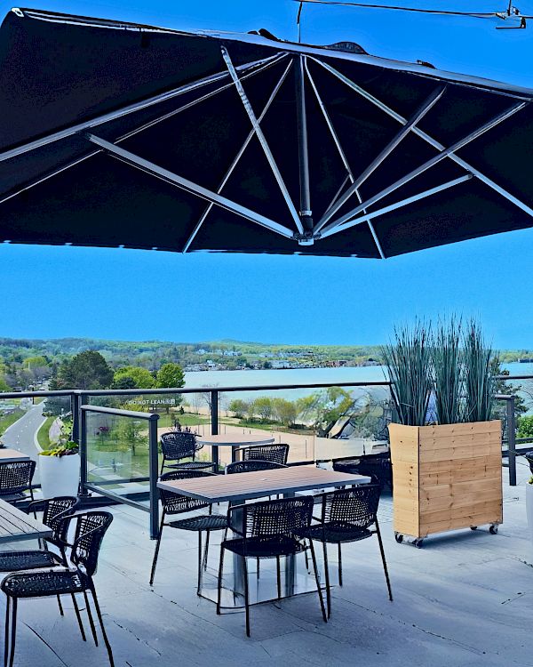 A rooftop patio with tables, chairs, a large umbrella, and scenic views, featuring modern outdoor furniture and clear blue skies.