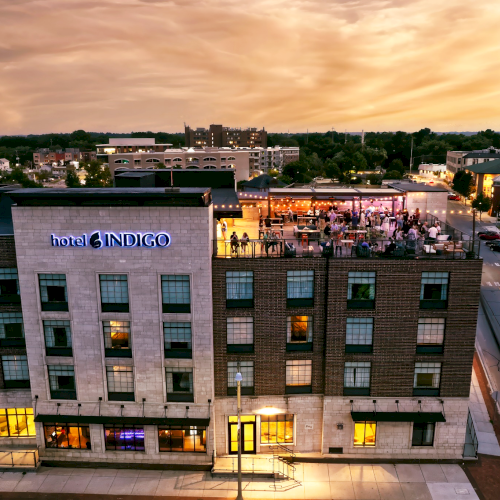 A hotel building with "Hotel Indigo" signage, featuring a rooftop bar with people, under a sunset sky.
