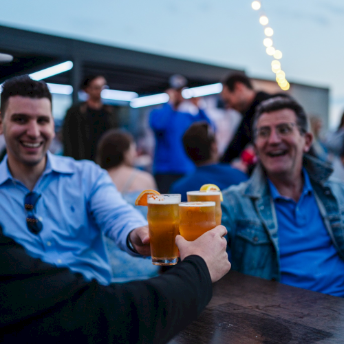 People enjoying drinks and toasting at an outdoor gathering, with string lights and a relaxed atmosphere in the background.