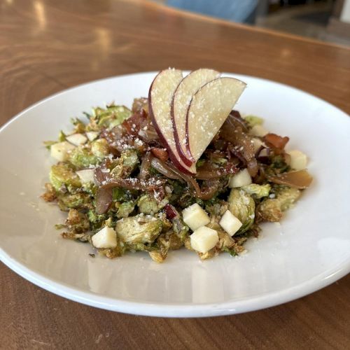 The image shows a salad on a white plate with shaved Brussels sprouts, diced cheese, bacon, and apple slices on top, placed on a wooden table.