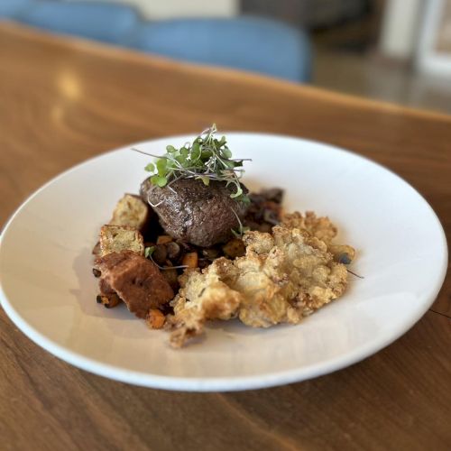 A plated dish features a piece of meat topped with greens, alongside crumbled and crispy elements, served on a white plate.