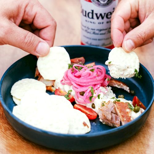 A dish with crackers, pickled onions, and meat is being served, accompanied by a Budweiser beer can.
