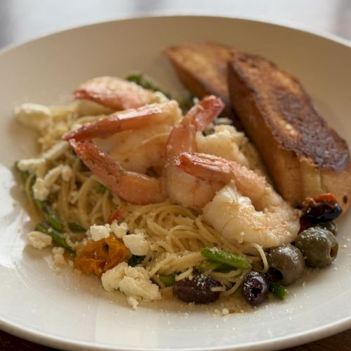 A dish with shrimp, pasta, olives, cherry tomatoes, and two pieces of toast on a white plate.