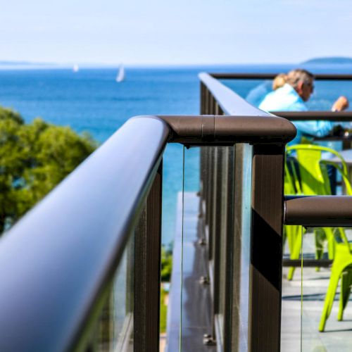 A terrace with people dining next to a glass railing overlooks a scenic ocean view with green chairs and distant trees visible.