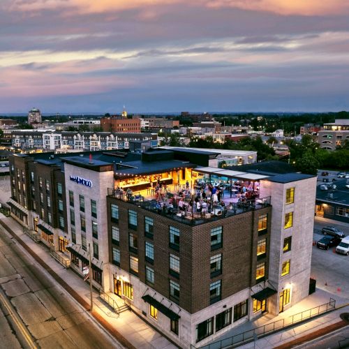 The image shows an aerial view of a modern building with a rooftop bar, surrounded by city streets and other buildings at dusk.