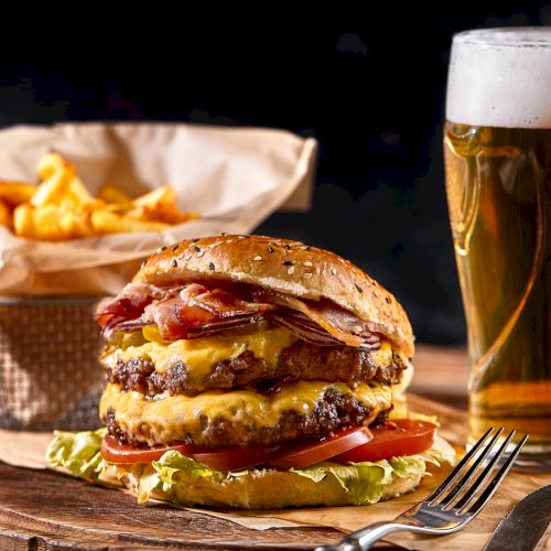 A burger with bacon and cheese on a wooden board, alongside fries in a basket and a glass of beer, on a rustic wooden table.