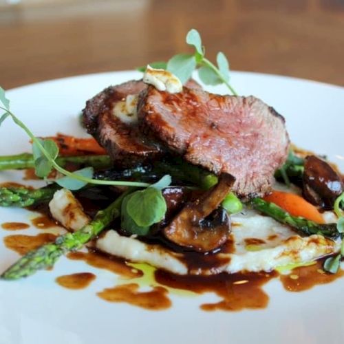 A gourmet dish featuring sliced meat on a bed of mashed potatoes, topped with asparagus, mushrooms, and herbs, drizzled with sauce.