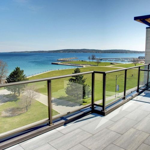 A balcony with glass railing overlooks a grassy park and a scenic body of water, with a clear blue sky in the background.