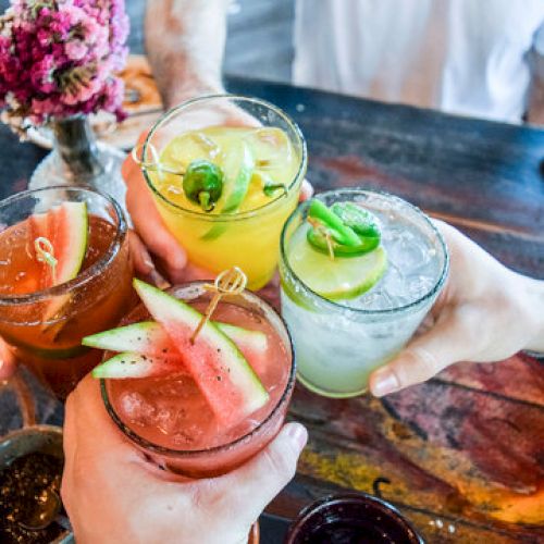 Four people are toasting with colorful cocktails garnished with fruit, over a decorative table with a flower vase.