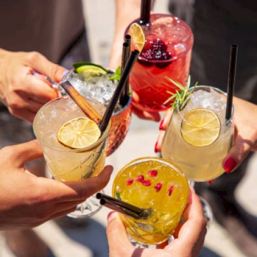 Five hands holding colorful cocktails with garnishes like lime slices, berries, and herbs, along with straws in each glass.