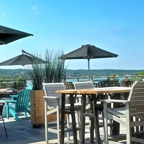 The image shows an outdoor patio with tables, chairs, and umbrellas, offering a scenic view of water and greenery under a clear blue sky.