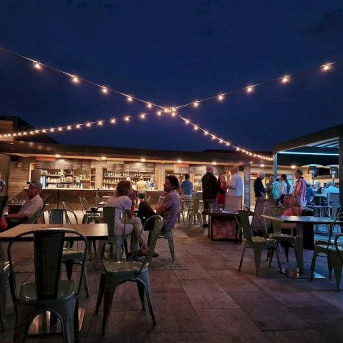 A rooftop bar at night with string lights, tables, and people socializing; a relaxed atmosphere.