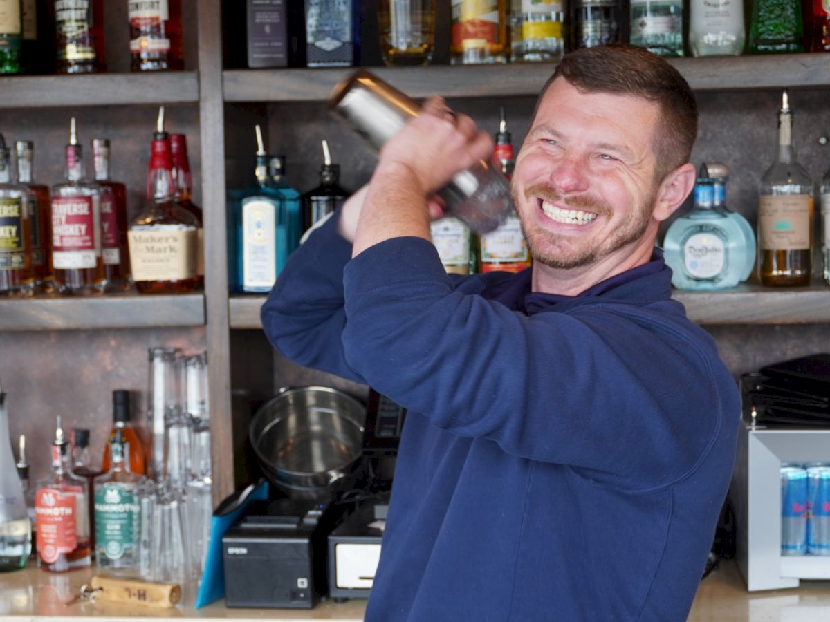 A person is smiling while shaking a cocktail shaker behind a bar stocked with various bottles and drinks.